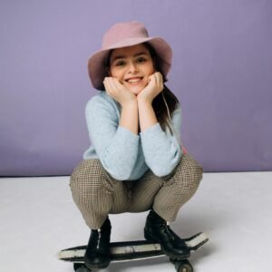 A cheerful teen girl smiling, crouching on a penny board, wearing a stylish hat indoors.