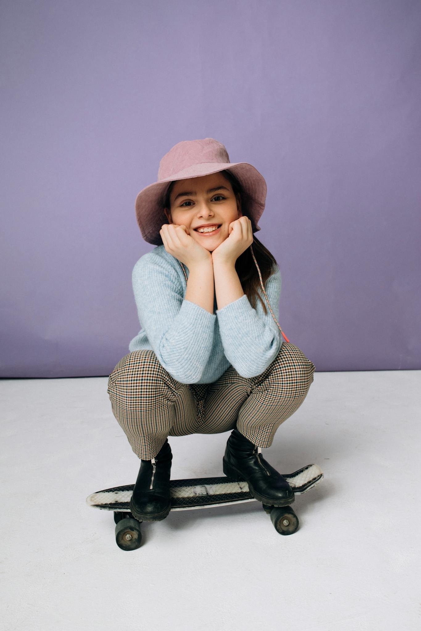 A cheerful teen girl smiling, crouching on a penny board, wearing a stylish hat indoors.