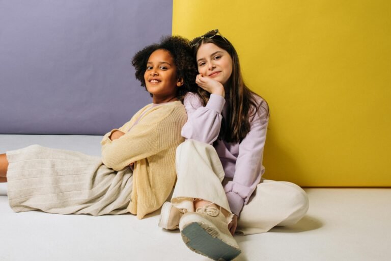Two young girls smiling and posing playfully against a vibrant yellow and purple backdrop. Stylish and cheerful vibe.