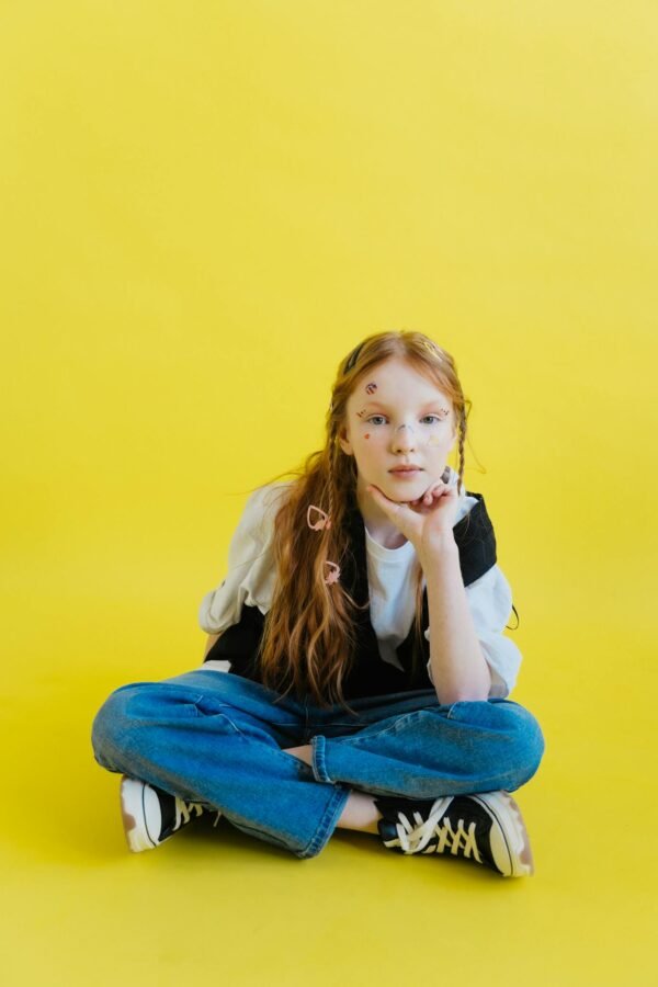 Young girl sitting casually on a vibrant yellow background with fun facial stickers.